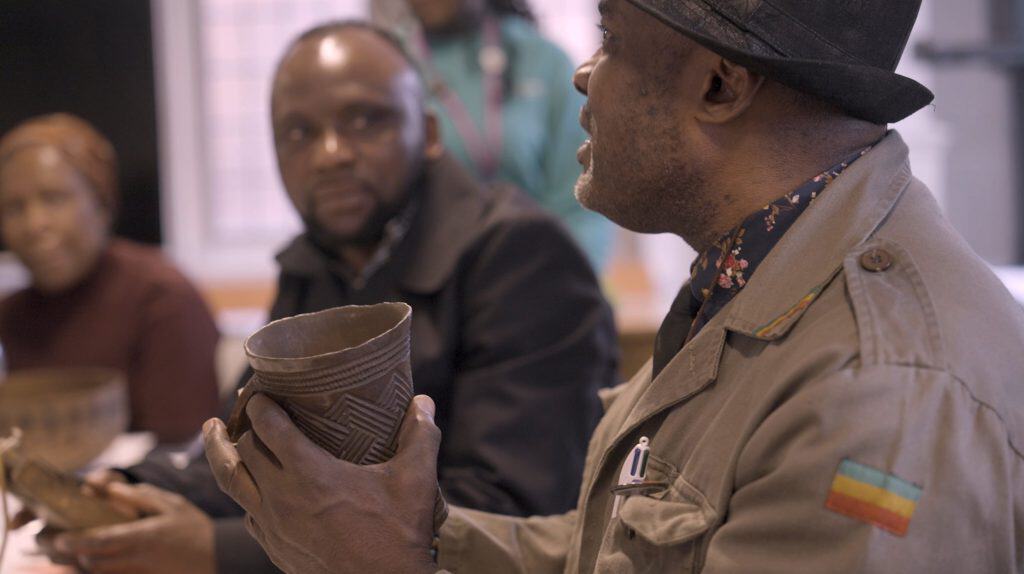 A man of African heritage holds a wooden cup in his hand while talking to other people.