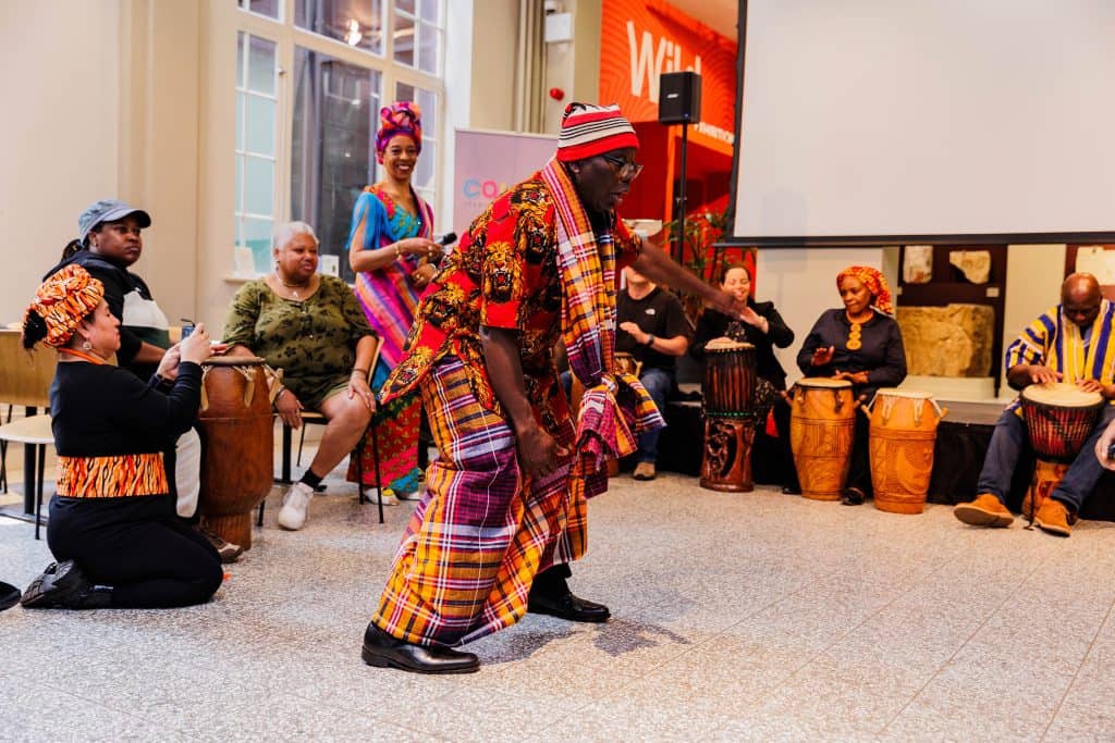 A man in traditional African dress dances in then Main Hall at Manchester Museum, surrounded by people in African dress playing drums.